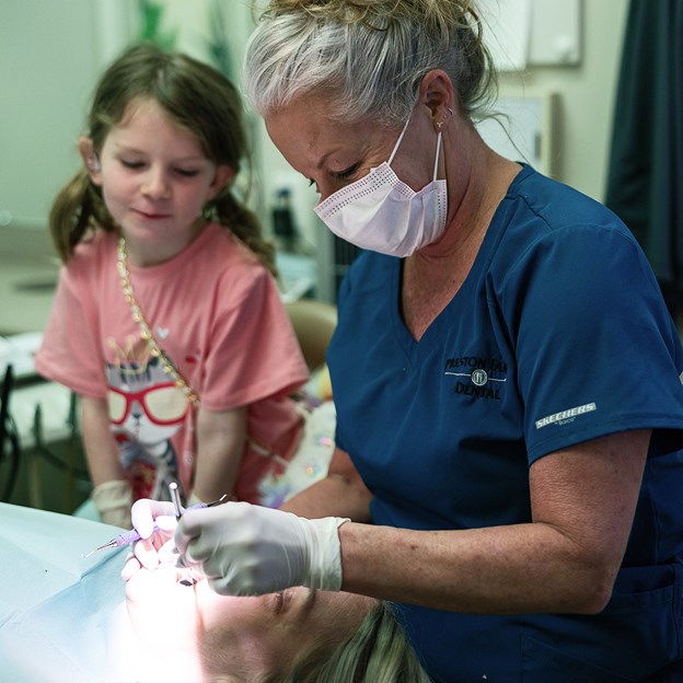 Dentist performing a dental exam on a patient while a young girl watches